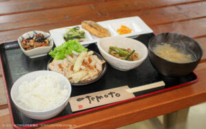 黒いトレーの上に豚肉しょうが焼き、にんにくの芽とひき肉の炒め物、タコの酢味噌和え、サバのから揚げ、ひじきの煮物、漬物、ごはん、みそ汁が並んでいる。