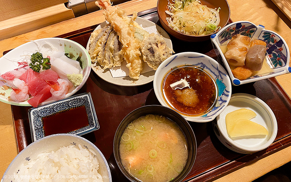 木製の盆に刺身、天ぷら、サラダ、煮物、漬物、みそ汁、白米が並ぶ。